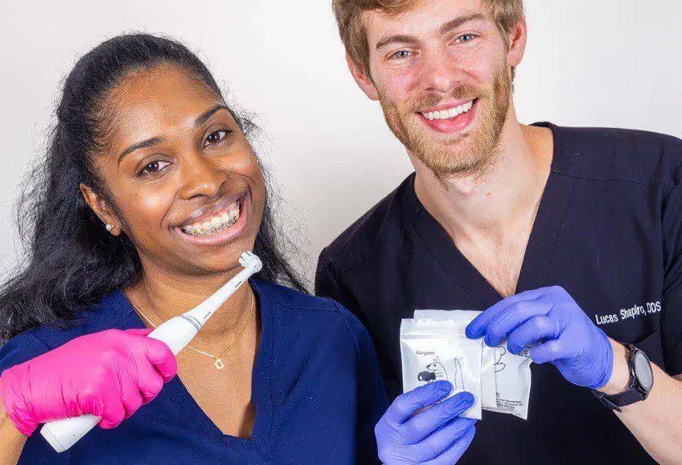 A Wall Street Orthodontics team member wearing braces and holding an electric toothbrush, beside Dr. Lucas Shapiro holding a packet of orthodontic elastics