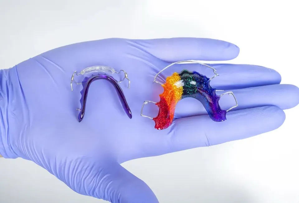 A gloved hand holding two removable retainers, one clear and one with a rainbow-coloured acrylic body